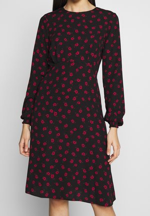 Zwarte knielange jurk met lange mouwen en rood bloemenpatroon, gedragen door een persoon met ontspannen handen langs het lichaam.