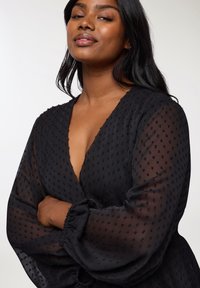 Czarna, przezroczysta bluzka z głębokim dekoltem w kształcie litery V, teksturowanym wzorem w kropki i bufiastymi rękawami. Lekka tkanina z elastycznymi mankietami.