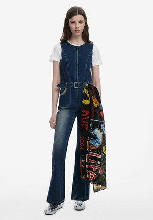 Jeans-Overall mit taillierter Taille, weitem Bein und einem Gürtel. Kombiniert mit einem weißen T-Shirt und einem bunten, gemusterten Schal, der über den Arm drapiert ist.