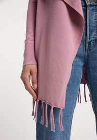 Scialle lavorato a maglia rosa con polsini a coste, bordi frangiati e una texture morbida, indossato sopra dei jeans in denim blu.