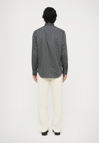 Czarna koszula z długim rękawem z białym, fakturowanym wzorem, z kołnierzykiem, noszona z beżowymi spodniami. Koszula ma prosty dół i mankiety na guziki.