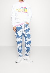 Felpa con cappuccio bianca con logo "The North Face" multicolore, abbinata a jeans blu tie-dye e sneakers bianche, con dettaglio a catena sulla cintura.