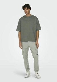 Graues, oversized T-Shirt mit kurzen Ärmeln, kombiniert mit hellgrauen Cargo-Hosen, die seitliche Taschen und tapered Cuffs haben. Weiße Sneaker runden den Look ab.