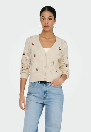 Vrouw draagt een crèmekleurige gebreide cardigan met geborduurde kersen, lichtblauwe jeans met hoge taille, creoolse oorbellen en een wit topje eronder.