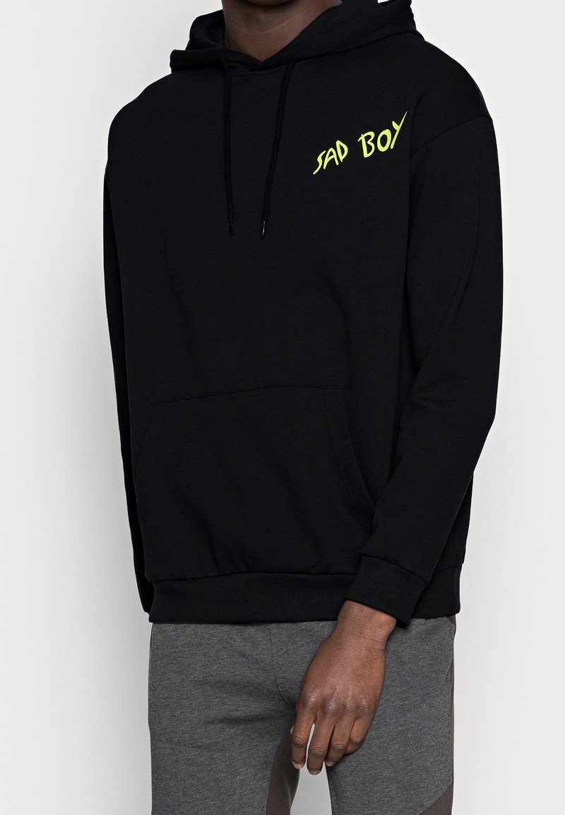 Il hoodie nero presenta una tasca frontale e un cappuccio con coulisse. Testo "SAD BOY" in giallo neon sul petto. Tessuto morbido, polsini a coste.