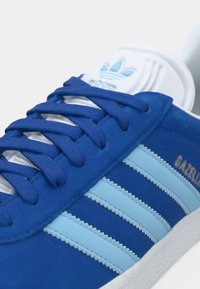 Μπλε σουέτ αθλητικό παπούτσι με ανοιχτές μπλε ριγέ λεπτομέρειες, λευκά κορδόνια και υφασμένο πέλμα. Διαθέτει χρυσό λογότυπο "GAZELLE" στο πλάι.