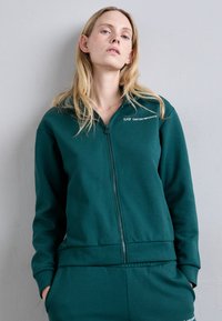 Πράσινη zip-up ζακέτα φτιαγμένη από μαλακό ύφασμα, με όρθιο γιακά, ριμπ μανσέτες και το λογότυπο "EA7 EMPORIO ARMANI" στο στήθος.