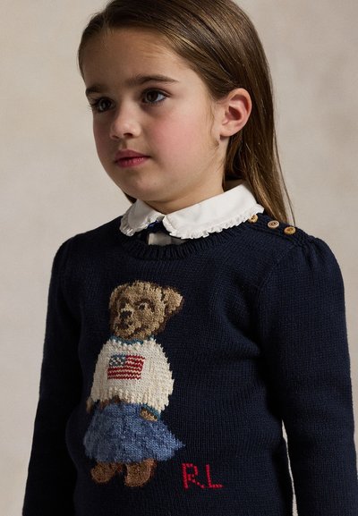 Junges Mädchen mit glatten braunen Haaren, das einen dunkelblauen Pullover mit einem Teddybär und ein weißes Hemd mit Rüschenkragen trägt, schaut leicht zur Seite.