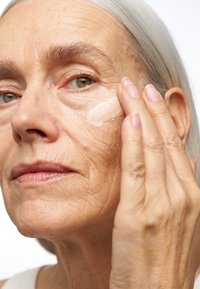 Een persoon met lang zilverachtig haar brengt een crème aan op hun wang, waarbij de gestructureerde huid en zichtbare rimpels worden benadrukt, in een close-up opmaak.