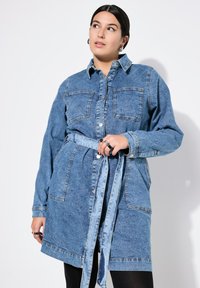 Vestido de camisa de mezclilla en azul claro, con dos bolsillos en el pecho, mangas largas y un cinturón para anudar. Textura suave con acentos de mezclilla en contraste.