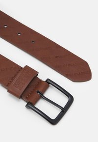 Cintura in pelle marrone con motivo impresso "Urban Classics", dotata di fibbia in metallo nero e superficie liscia. Le estremità sono affusolate.