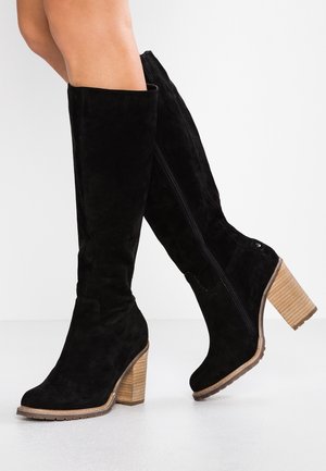 Bottes hautes noires en suède avec talons blocs en bois, fermeture éclair latérale, portées par une personne debout sur un sol clair.