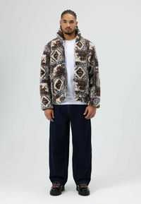 Pluizige jas met een geometrisch patroon in bruin, blauw en crème, met een hoge kraag en een open voorkant, gecombineerd met donkerblauwe jeans met wijde pijpen en zwarte schoenen.