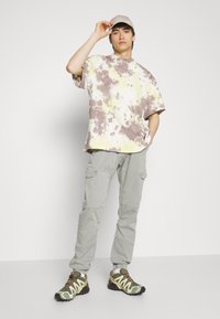 Tânăr îmbrăcat cu un tricou tie-dye, pantaloni cargo gri deschis, pantofi de drum verzi și o șapcă bej, pozând pe un fundal alb.