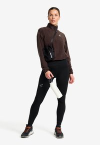Brauner Fleece-Pullover mit halbem Reißverschluss, schwarze Leggings, hält eine weiße Wasserflasche. Eine kleine schwarze Tasche mit einem Gurt hängt von der Schulter.