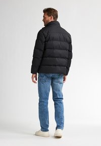 Uomo che indossa un giubbotto imbottito nero, jeans blu e sneakers bianche, in piedi e che guarda di lato su uno sfondo bianco.