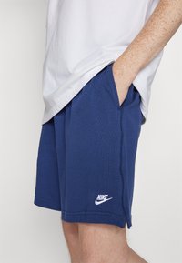 Pantaloni sportivi blu navy con tasche laterali, texture morbida e logo Nike bianco sull'orlo inferiore. Include dettagli di cucitura visibili.