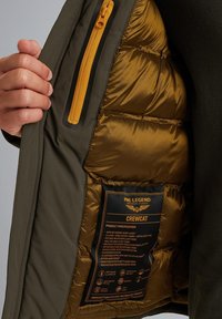 Oliven grøn ripstop kort jakke med en gylden puffy-foring, der har en gul lynlåslomme og et mærke med produktbeskrivelser.