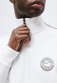 Homme portant un pull zippé blanc avec le logo "Boss Tennis Club 1997", main tirant la fermeture éclair près du col, gros plan sur le haut du torse et le visage.