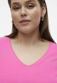 Roza v-neck majica s gladko teksturo, ki ima kratke rokave. Model nosi zlate uhane in ima srednje dolge lase.