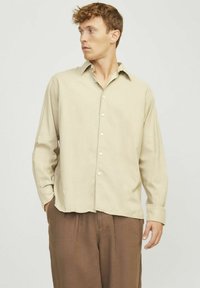 Helles beige Hemd mit Knöpfen und spitzem Kragen, langen Ärmeln und lockerer Passform. Kombiniert mit braunen Leinenhosen.