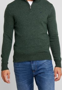 Mörkgrön zip-up tröja, strukturerad stickad tyg, figursydd design, ribbade mudd och nederkant, liten logotyp på bröstet, kombinerad med blå jeans.