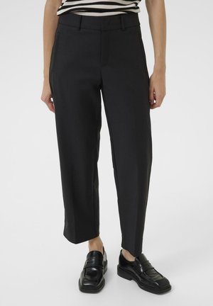Persoon die zwarte cropped rechte broek en zwarte loafers draagt, met een deel van een zwart-wit gestreept topje zichtbaar boven de taille.
