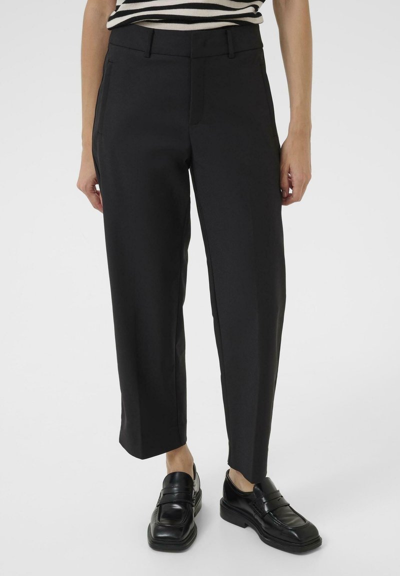 Personne portant un pantalon noir court à jambes droites et des mocassins noirs, avec une partie d'un haut rayé noir et blanc visible au-dessus de la taille.