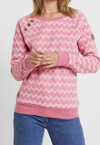 Felpa rosa con motivo a zigzag in una tonalità di rosa più chiara, polsini a coste e bottoni decorativi sulla spalla. Realizzata in tessuto morbido.