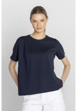 Kobieta ubrana w jednolitą granatową koszulkę z krótkim rękawem i białe spodnie, stoi z jedną ręką w kieszeni na tle jasnego, jednolitego tła.