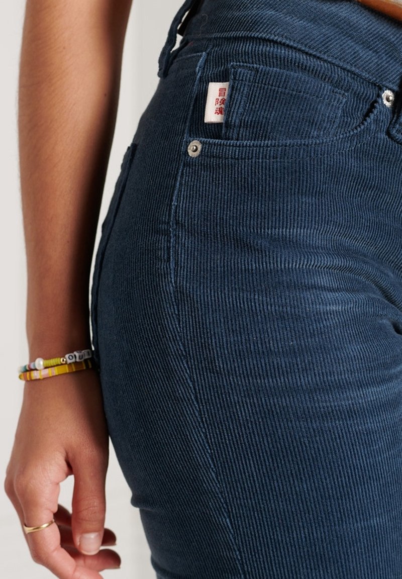 Jeans de pana azul oscuro con canalones verticales, que presentan una etiqueta de parche en la cinturilla y un pequeño detalle de botón plateado.