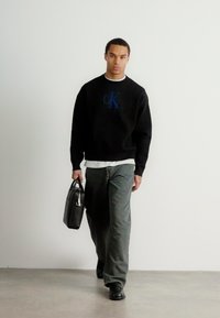 Čierna mikina s modrým logom CK, sivé nohavice a čierne topánky. Drží textúrovanú čiernu kabelku. Jednoduchý, moderný dizajn, minimalistické detaily.