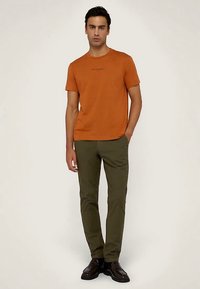 Pomarańczowa koszulka z krótkim rękawem i okrągłym dekoltem, z delikatnym nadrukiem na piersi, w połączeniu z oliwkowozielonymi spodniami o wąskim kroju i ciemnymi butami.