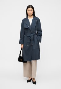 Μπλε ναυτικό παλτό από αδιάβροχο υλικό, με ζώνη στη μέση, μεγάλους γιακάδες και ασημένια εξαρτήματα. Συνδυασμένο με μπεζ παντελόνια φαρδιάς γραμμής.