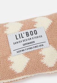 Adīts bēšs un persiku spilvens ar dimanta rakstu. Uz tā ir balta birka ar melnu tekstu, kurā teikts "LIL'BOO" un dizaina detaļas.