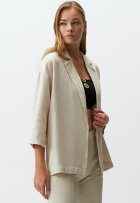 Hellbeige Leinenblazer mit entspannter Passform, Schalkragen, Dreiviertelärmeln und Seitentaschen, kombiniert mit einem schwarzen Crop-Top.