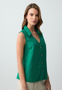 Blusa verde senza maniche con colletto a giro, dotata di chiusura a bottoni e realizzata in un tessuto leggero e testurizzato. Vestibilità leggermente rilassata.