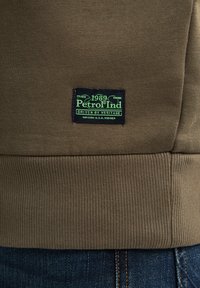 Olivově zelená látka na mikinu s žebrovaným lemem. Obsahuje černou samolepku se zeleným textem, na které je napsáno "Petrol Ind 1989 Driven by Heritage."