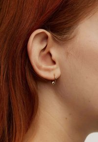 Pendiente de aro de oro con una superficie suave y pulida y un pequeño detalle de nudo adjunto. Lleva puesto en la oreja de un modelo, mostrando la forma y el diseño.