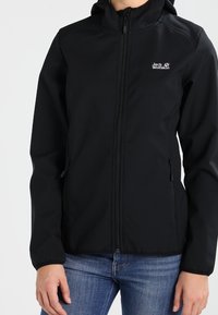 Veste en softshell noire avec capuche, dotée d'une fermeture éclair frontale, de poches latérales et d'un logo sur le côté gauche de la poitrine. Texture lisse et design ajusté.