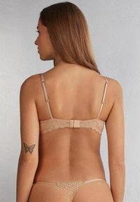 Spets-bh och string-set i nude-färg, med justerbara band, en dekorativ bakknäppning och intrikata spetsdetaljer längs kanterna.