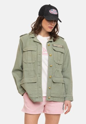 Femme portant une veste utilitaire verte avec des boutons dorés et un écusson, un t-shirt blanc imprimé, un short rose et une casquette noire avec un logo coloré.