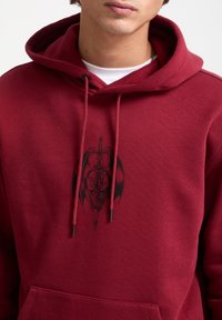 Sudadera roja hecha de un tejido suave, con un diseño gráfico negro de una calavera con una espada y alas. Incluye cordones y un bolsillo en la parte frontal.