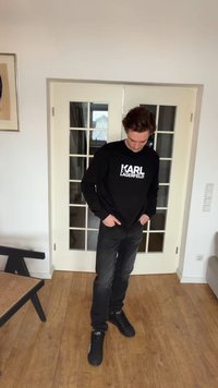 Άνδρας που φοράει μαύρο φούτερ Karl Lagerfeld, μαύρο τζιν και μαύρα αθλητικά παπούτσια, στέκεται μέσα σε ξύλινο δάπεδο, με τα χέρια στις τσέπες, κοιτάζοντας προς τα κάτω.