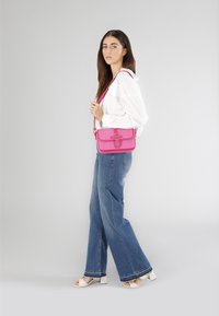 Lyserød crossbody taske med et struktureret stof, flaplukning og dekorativ hardware. Accentueret med en bøjet rem. Båret med lyse denimbukser.