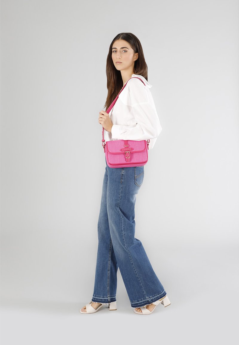 Lyserød crossbody taske med et struktureret stof, flaplukning og dekorativ hardware. Accentueret med en bøjet rem. Båret med lyse denimbukser.