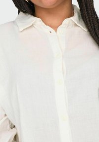 Chemise blanche légère à boutons, en tissu doux et texturé, avec un col classique. Détails de couture subtils et bords légèrement arrondis.