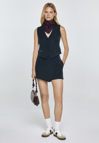Gilet a righe blu navy abbinato a pantaloni corti, con tasche laterali doppie. Abbinato a una sciarpa a fantasia viola, calzini bianchi e sneakers multicolori.