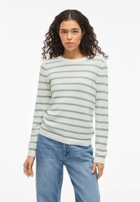 Femme aux cheveux bouclés portant un pull en tricot blanc à rayures vertes et un jean bleu, debout sur un fond blanc.