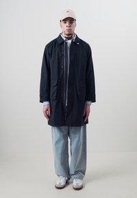 Navy long coat with button closure and side pockets, layered over a light shirt. Paired with loose light denim pants and white sneakers.